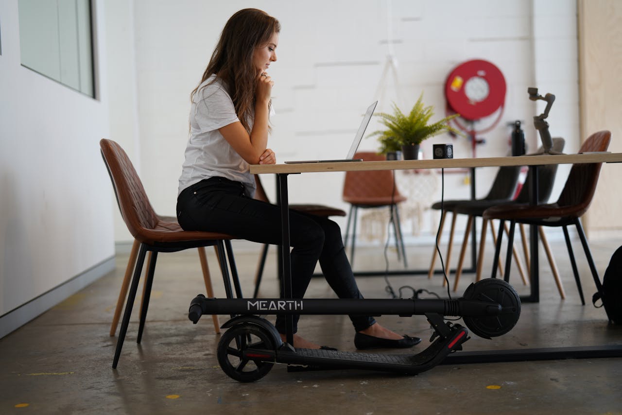 A woman sits at a table indoors working on a laptop with an electric scooter nearby, showcasing a modern lifestyle.